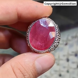 May include: A large, oval-cut ruby gemstone ring. The ruby is a deep red color and is set in a silver-colored band with a detailed, textured design around the stone. The ring is held by a person's hand.