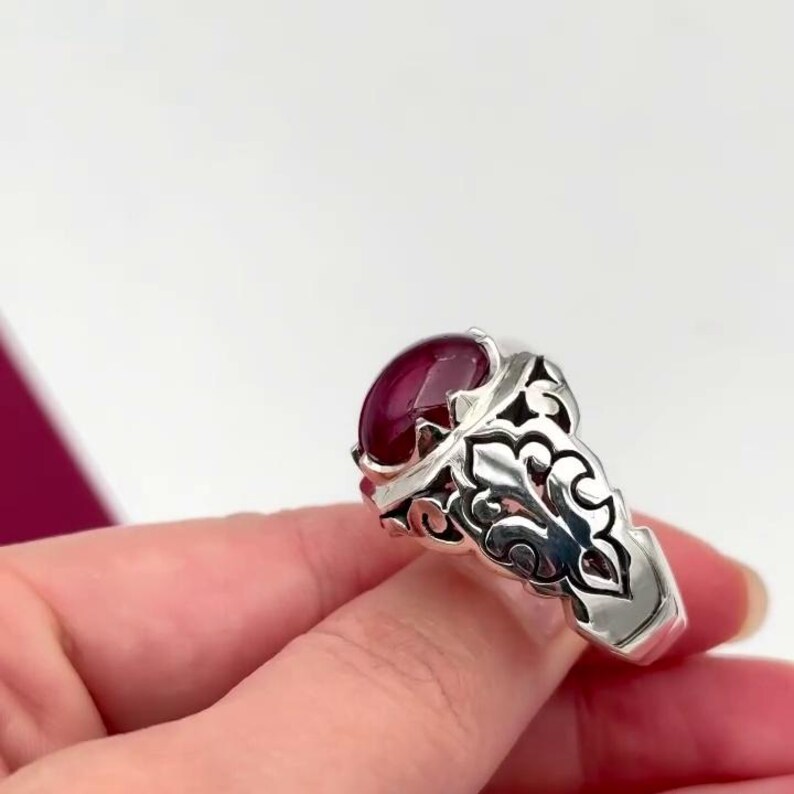 May include: A silver ring with an ornate design and a large, oval-shaped, dark red gemstone.