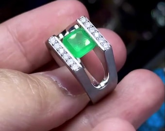 May include: A silver ring featuring a square, vibrant green gemstone at its center. The ring's band has a modern design with a row of small, clear gemstones on each side of the central stone. The ring is held in a hand.