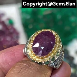 May include: A silver and gold ring featuring a large, faceted, oval-shaped, deep red ruby gemstone. The ring has a detailed design around the gemstone and on the band. The text "Copyright@GemsElan" is visible at the top of the image.