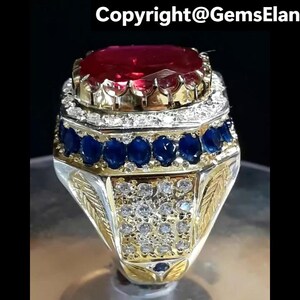 Könnte beinhalten: Ein aufwendiger Ring mit einem leuchtend roten Edelstein. Der Ring hat ein Gold- und Silberband mit einer Reihe blauer Edelsteine und kleineren weißen Steinen. Das Design weist detaillierte Muster und eine polierte Oberfläche auf.