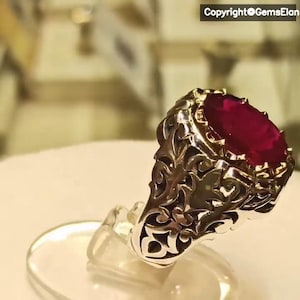 May include: A silver ring with an ornate design features a large, oval-shaped ruby gemstone. The ring is set on a white display stand.