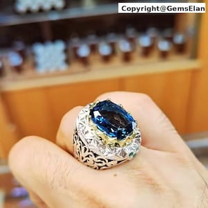 Könnte beinhalten: Ein aufwendiger Ring mit einem großen, oval geschliffenen, tiefblauen Edelstein. Der Ring hat ein silbernes Band mit detaillierten Filigranarbeiten und goldenen Akzenten. Der Text "Copyright@GemsElan" ist sichtbar.