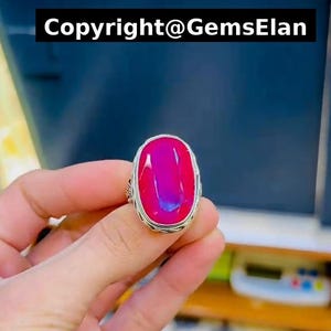 May include: A silver ring featuring a large, oval-shaped, vibrant pink cabochon gemstone with a smooth, polished surface. The metal band displays intricate, textured detailing along the edges. The text Copyright@GemsElan is visible at the top.