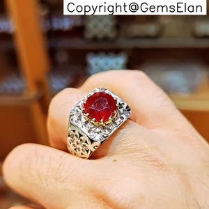 Könnte beinhalten: Ein silberner Ring mit einem großen, facettierten, roten Edelstein. Der Ring hat eine quadratische Fassung mit einem floralen Design an den Seiten. Der Edelstein ist in einer goldfarbenen Lünette gefasst.