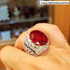 Op de afbeelding: Een zilveren ring met een grote, ovale robijn-edelsteen gezet in een gedetailleerd, versierd ontwerp. De ring zit aan een vinger.