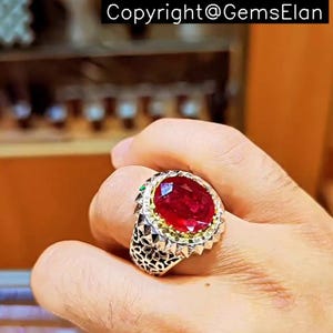 May include: A silver ring with a large, oval-shaped ruby gemstone set in a gold-toned bezel. The ring has a detailed, textured band with a decorative design.