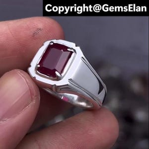May include: A silver ring with a rectangular, deep red gemstone. The ring has a polished, reflective surface and a classic, masculine design. The text "Copyright@GemsElan" is visible at the top.