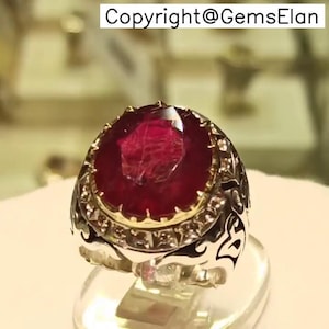 May include: A silver ring with an ornate design features a large, oval-shaped ruby gemstone surrounded by smaller, white gemstones.