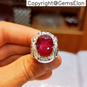 May include: A silver ring with a large, oval-cut ruby gemstone. The ruby is a deep red color and is set in a gold-colored bezel. The ring has intricate silver detailing around the gemstone.