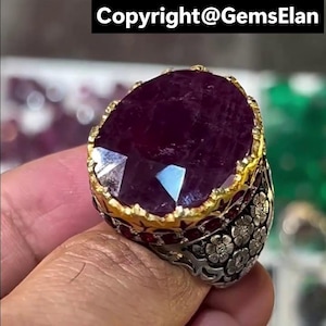 Könnte beinhalten: Ring mit einem oval geschliffenen Rubin, einer goldfarbenen Lünette und einem silberfarbenen Band. Das Band hat ein Blumenmuster. Der Ring wird von den Fingern einer Person gehalten. Der Text "Copyright@GemsElan" steht oben.