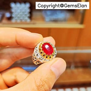 May include: A silver ring featuring a large, oval red gemstone. The ring has a gold setting around the gemstone and detailed silver patterns on the band. The image includes the text "Copyright@GemsElan".