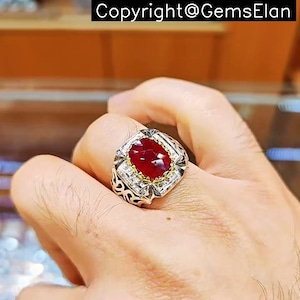 May include: A silver ring featuring a large, oval-shaped red gemstone set in a gold-toned bezel. The ring has a detailed, intricate design with smaller white gemstones surrounding the main stone.