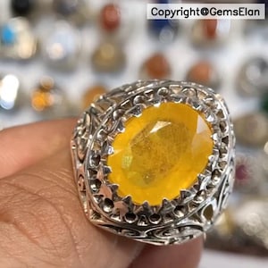May include: A silver ring featuring an oval, yellow gemstone. The ring has an intricate, filigree pattern around the stone and on the band. The gemstone is a vibrant, golden yellow. The background is blurred, with other jewellery visible.