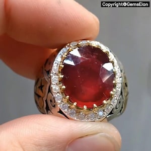 May include: A silver ring with a large, faceted, deep red gemstone at the center. The gemstone is surrounded by a halo of small, clear stones. The ring's band features an ornate, engraved design. The text "Copyright@GemsElan" is visible.
