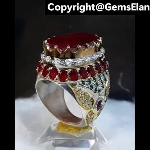 Op de afbeelding: Een meerlagige ring met een grote, ovale, dieprode edelsteen aan de bovenkant. De ring heeft meerdere banden van kleinere rode en groene edelstenen en heldere stenen. De ring heeft een goud- en zilverkleurig metalen ontwerp.