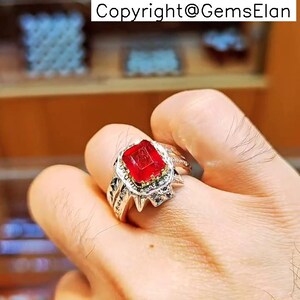 May include: A silver ring featuring a large, square-cut, bright red gemstone. The ring has intricate detailing around the gemstone and along the band. The image includes the text "Copyright@GemsElan".