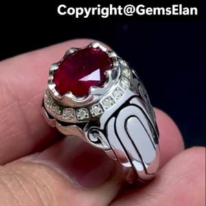 May include: A silver ring with a large, oval-cut ruby gemstone. The ring features a decorative design with small, clear accent stones around the ruby. The ring's band has a detailed, ornate pattern.