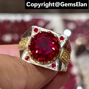 Peut inclure: Une bague en argent et or avec une grosse pierre précieuse rouge foncé facettée en son centre. De petites pierres rouges sont serties autour de la pierre principale et sur les épaules de la bague. La bague a un design rectangulaire avec des accents dorés et le texte "Copyright@GemsElan".