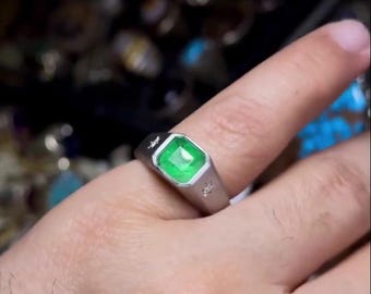 May include: A silver ring with a square, emerald-green gemstone. The ring has a geometric design with a star and other small details. The ring is on a finger.