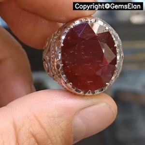 Peut inclure: Une bague en argent ornée d'une pierre précieuse rouge foncé, ovale et facettée. Le chaton de la bague présente un motif ornemental texturé. L'image contient le texte "Copyright@GemsElan".