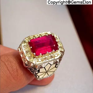 May include: A gold and silver ring with a large, red, emerald-cut gemstone surrounded by smaller, clear gemstones.