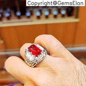May include: A silver ring featuring a large, rectangular, vibrant red gemstone. The ring is surrounded by smaller, clear stones and has an ornate, detailed band. The text "Copyright@GemsElan" is visible at the top.