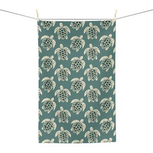 May include: A rectangular tea towel featuring a repeating pattern of off-white sea turtles printed on a solid teal background. The turtles are arranged in a grid-like formation across the entire surface of the fabric, which hangs from a thin black line.