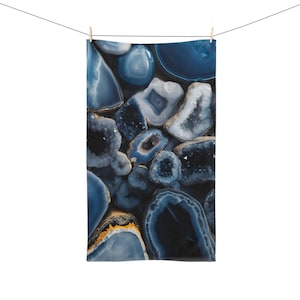 May include: A rectangular towel featuring a pattern of blue and white agate stone slices. The towel is hanging from a clothesline with a wooden clothespin. The agate slices have a variety of shapes and sizes, with some having a dark blue center and white edges.