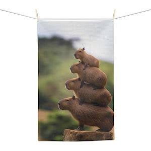 May include: A white waffle-textured tea towel hanging from a clothesline, featuring a photographic print of four brown capybaras stacked vertically on top of each other, with the bottom capybara standing on a wooden stump in a grassy field.