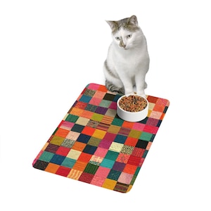 May include: A rectangular pet mat featuring a vibrant, multi-colored patchwork grid pattern of small squares in shades of red, orange, yellow, green, blue, and black. A white bowl filled with dry brown pet food sits on the mat's surface.