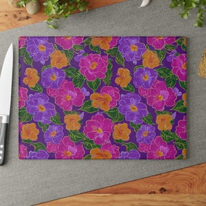 Pode incluir: Tábua de corte retangular com um padrão floral vibrante. O design apresenta flores roxas, rosas e laranjas com folhas verdes sobre um fundo roxo escuro. A tábua é feita de um material liso e durável.