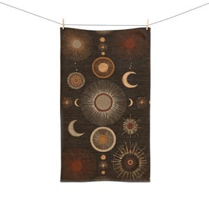 May include: A dark brown towel with a celestial design featuring suns, moons, and circular patterns in shades of beige and brown. The towel is hanging from a clothesline with a wooden clothespin.