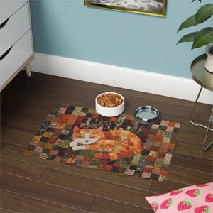 May include: A rectangular pet mat featuring a patchwork design of various brown, orange, and green fabric squares. A stylized orange cat with a white face and closed eyes is centered on the mat. A white bowl of dry food and a silver bowl sit on the mat.