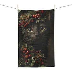 May include: A rectangular kitchen towel featuring a detailed print of a black cat surrounded by festive holly leaves and bright red berries. The cat has large, expressive eyes and is framed by greenery at the top and bottom of the fabric surface.