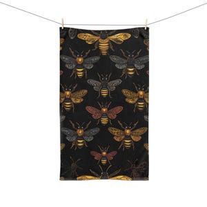 Pode incluir: Uma toalha retangular com um padrão repetitivo de abelhas sobre um fundo preto. As abelhas são representadas em vários tons de amarelo, laranja e cinza, cada uma exibindo designs decorativos intrincados nas asas e corpos listrados.