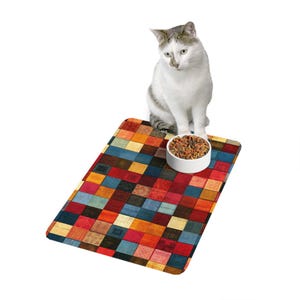 Puede incluir: Un tapete rectangular para mascotas con un patrón de cuadrícula multicolor en tonos rojo, azul, naranja y amarillo. Un cuenco de cerámica blanco lleno de comida seca marrón descansa sobre el tapete, junto a un gato de color blanco y gris.