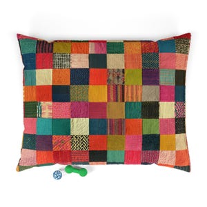May include: A large rectangular pillow featuring a patchwork design of various multicolored fabric squares in shades of red, blue, green, yellow, pink, and orange. A small blue spherical toy and a green dumbbell-shaped toy are positioned below.
