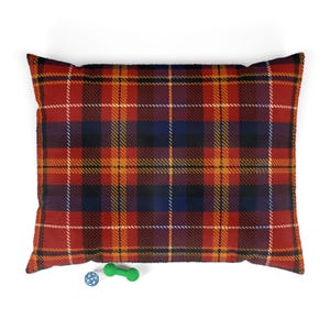 May include: A rectangular pet bed featuring a vibrant red, navy blue, and yellow plaid tartan pattern. The fabric has a soft, textured appearance. Positioned below the bed are a small blue spherical toy and a green dumbbell-shaped toy.
