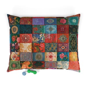 May include: A square patchwork pillow featuring a variety of colorful fabric squares with diverse floral, geometric, and ornate patterns in shades of red, blue, green, orange, and yellow. A small blue ball and a green dumbbell-shaped toy are below.