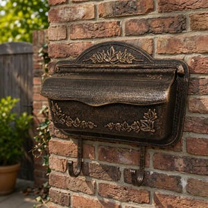 May include: A wall-mounted mailbox with a dark bronze finish and a weathered texture. The rectangular box features a curved top lid and two bottom hooks. Embossed leaf motifs decorate the top arch and the lower corners of the front panel.