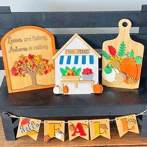 May include: Wooden fall-themed decorations on a black bench. Includes a sign with a tree and text, a farm stand, and a cutting board with pumpkins. A banner spells "FALL".