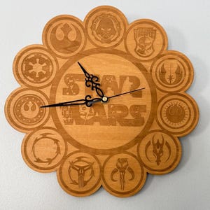 May include: Wooden Star Wars themed clock with a flower-like design. The clock features various Star Wars symbols and the words "STAR WARS" in the center. The clock hands are black, and the clock is made of light brown wood.