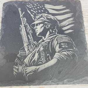 May include: A square, dark gray slate coaster featuring an etched image of a soldier holding a rifle, with an American flag in the background. The soldier is wearing a military uniform and cap. The design is detailed and patriotic.