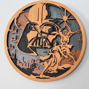 May include: A round wooden clock featuring a layered design with a Star Wars theme. The clock face includes Darth Vader's silhouette, figures with lightsabers, and a cityscape. The clock hands are black.