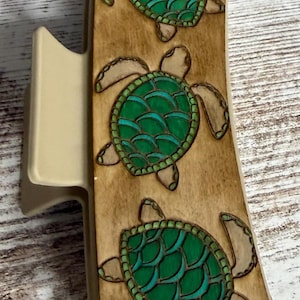 May include: Wooden hair clip featuring three green sea turtles with detailed shell patterns. The turtles are set against a light brown background, with a beige clip mechanism. The clip is approximately 4 inches long.