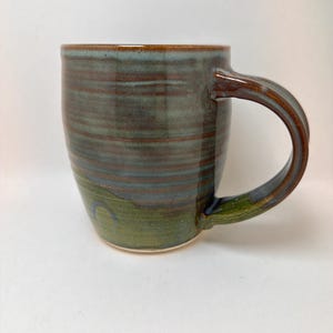 May include: A handcrafted ceramic mug with a rounded body and a large handle. The mug features a blend of colours, including shades of blue, green, and brown. The interior and handle are a darker brown. The mug is suitable for hot or cold drinks.