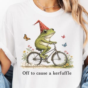 Könnte beinhalten: Weißes T-Shirt mit einer Illustration eines grünen Frosches auf einem Fahrrad, der einen rot-weiß gepunkteten Hut trägt. Der Text "Off to cause a kerfuffle" ist aufgedruckt. Schmetterlinge und Blumen ergänzen das Design. Ein lustiges Design.