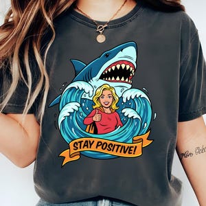 Könnte beinhalten: Dunkelgraues T-Shirt mit einem Cartoon-Hai, Wellen und einer Frau, die den Daumen hochhält. Das Shirt trägt den Text "STAY POSITIVE!" auf einem gelben Banner. Das T-Shirt wird mit einer blauen Jeans getragen.