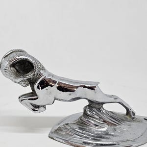 Pode incluir: Um adorno de capô de carneiro cromado para um carro. O carneiro está saltando para a frente com a cabeça virada para trás. O adorno é montado em uma base que se assemelha a uma rocha.